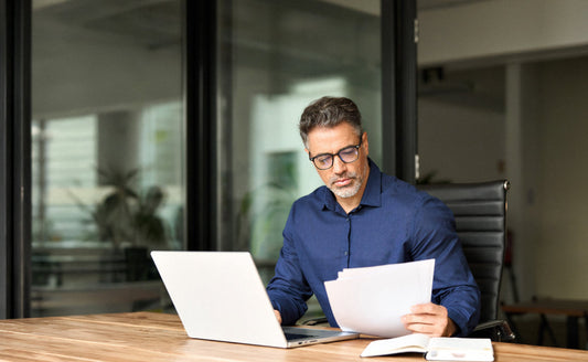 Middle-aged executive with glasses reviewing documents next to a laptop in a modern office.